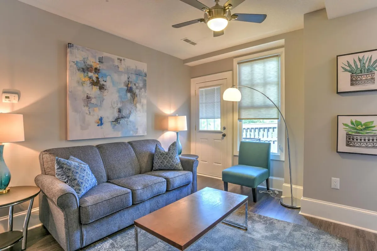 This image features a comfortable living room setting with a plush sofa, eye-catching artwork, a stylish coffee table, an inviting lamp, and a cozy chair. Natural light streams through large windows, highlighting the renovated, airy feel of the space. This arrangement captures both comfort and visual interest, emphasizing the room’s potential for both relaxation and social gatherings.