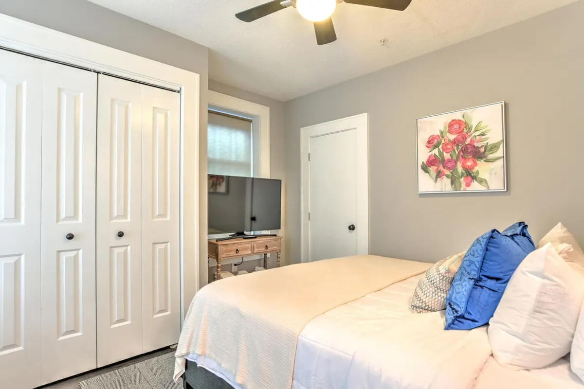 This image captures a tranquil bedroom with a comfortable bed adorned with decorative pillows, framed floral artwork on the wall, a television atop a wooden stand, and double closet doors. Gentle natural light fills the space, accentuating the serene and restful mood of the room.