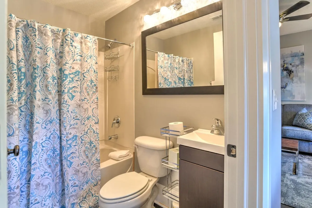 A bright, inviting bathroom featuring a blue-patterned shower curtain, a large vanity mirror, and modern fixtures. Morning light streams through the window, illuminating the space and creating a fresh, restful ambiance ideal for starting or ending the day.