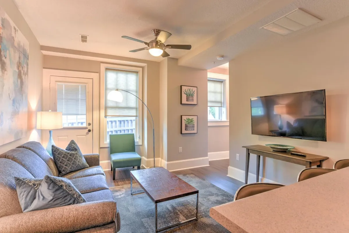 The image showcases a cozy, tastefully decorated living area featuring a plush sofa adorned with decorative pillows, a stylish green accent chair, a sleek coffee table ideal for casual gatherings, a modern television for entertainment, and eye-catching artwork that adds character to the space. The scene radiates comfort and invites relaxation, perfect for socializing, unwinding, or enjoying a quiet moment with a book or morning coffee.