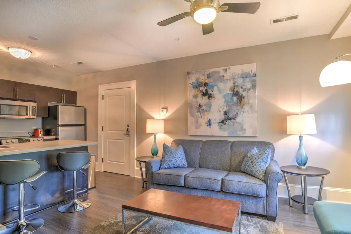 This view captures a different angle of the living area, including a comfortable sofa, modern lighting fixtures, and striking artwork, as well as a glimpse of the adjacent kitchen island. The interplay of soft and bright tones, along with contemporary touches, creates a welcoming, stylish environment suited for relaxation or socializing.