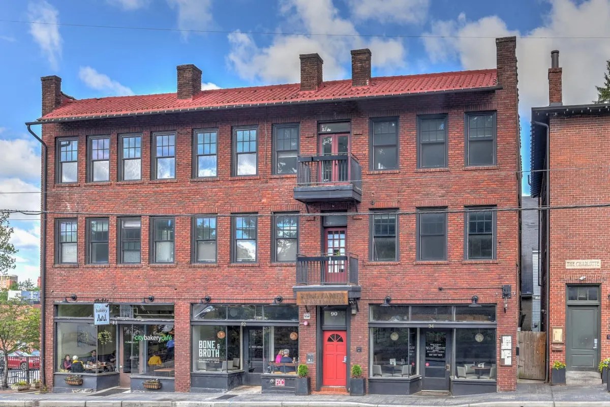 An exterior shot highlighting a red door, large windows, a classic brick facade, and a balcony. The scene conveys a welcoming atmosphere, with cues to social activities such as enjoying coffee, shopping, and participating in community events. The architecture and street view reinforce the area’s inviting urban vibe.