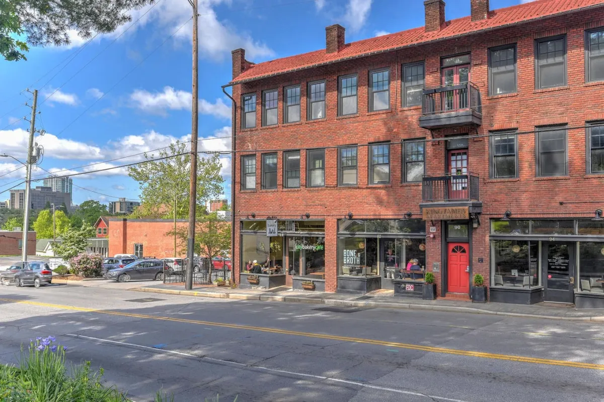 The image shows a brick building facade with balconies, storefronts, and surrounding greenery. The scene is active and urban, connecting the property to local dining and shopping experiences.
