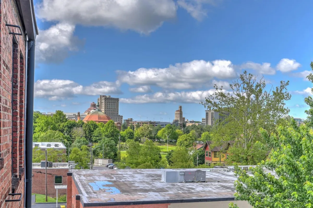 A wide view featuring a sky dotted with clouds, downtown buildings visible in the distance, and a green park area in the foreground. The atmosphere is serene and uplifting, with plenty of open space for walking, jogging, or attending outdoor events. The image hints at the proximity of downtown and the ease of walking to area attractions.