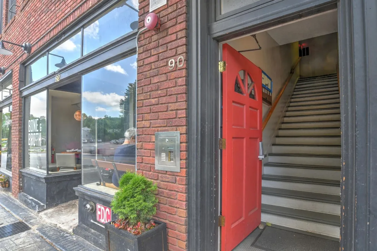 This image displays the red front door of the building, historic brick facade, a staircase leading up to the entry, large windows, and a welcoming planter box. The scene captures both the inviting entryway and hints at the historic charm of the architecture.