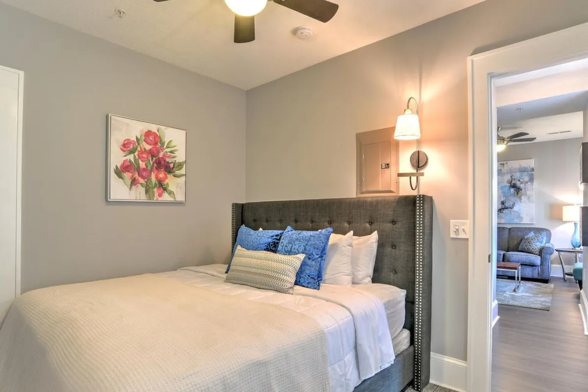 A serene bedroom scene featuring a comfortable bed adorned with decorative pillows, floral artwork above the headboard, and soft wall sconce lighting. The space is bathed in gentle natural light, creating a calming and inviting atmosphere perfect for relaxation after a day out.